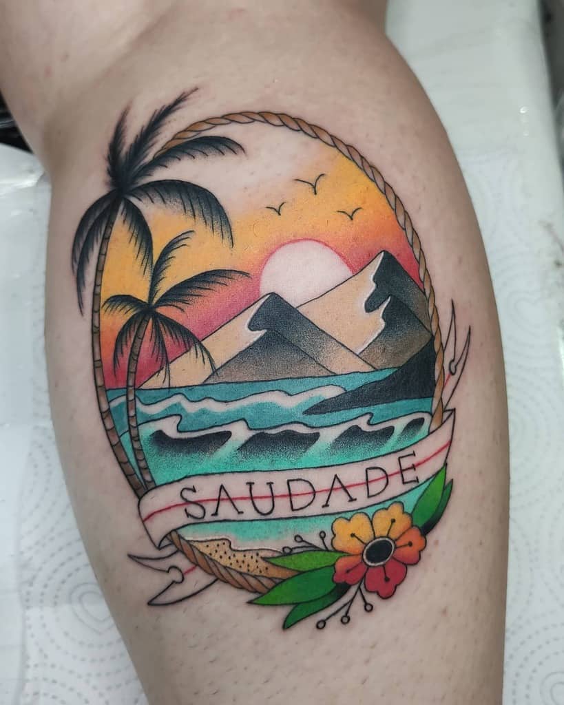 Colorful tropical tattoo featuring an ocean sunset, mountains, palm trees, and a banner with the word 'Saudade,' framed by rope and flowers