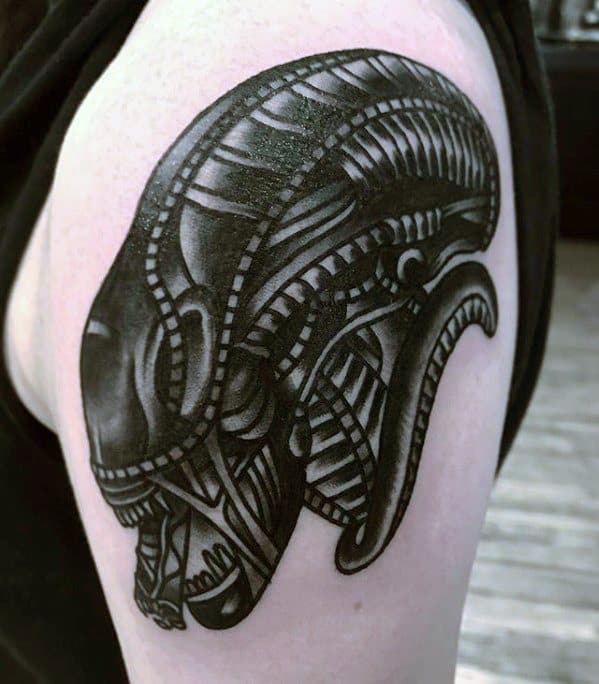 Bold black Xenomorph tattoo on upper arm with a tribal-inspired design, featuring thick lines and stylized alien features.