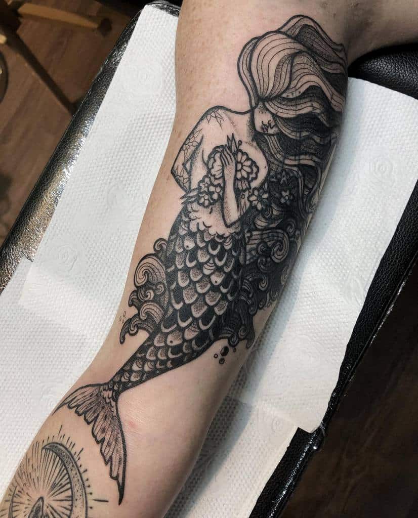 Black and grey tattoo of a mermaid with flowing hair, holding a seashell, with intricate scale details and swirling ocean elements.
