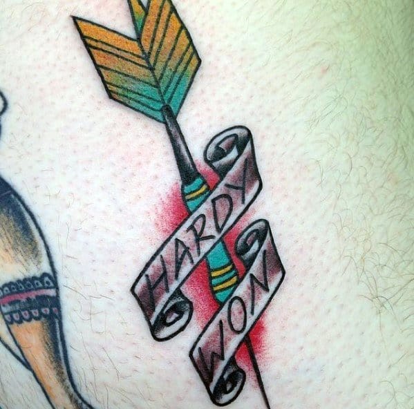 Traditional-style dart tattoo with green and yellow fletching, wrapped in a banner that reads “Hardy Won,” symbolizing victory through struggle.