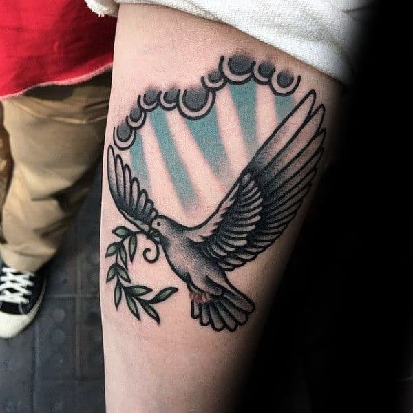 Tattoo of a flying dove with an olive branch surrounded by rays of light and clouds on an arm