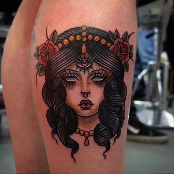 Tattoo of a mystical woman with a third eye, adorned with jewelry, flowers, and dark wavy hair, inked in bold colors on the thigh.