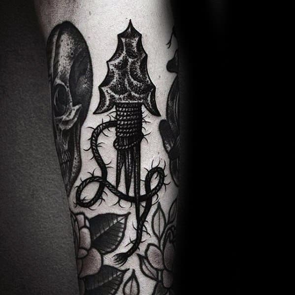 Dark blackwork broken arrow tattoo with a textured arrowhead and thorny vine wrapping around the shaft, surrounded by floral and skull elements.