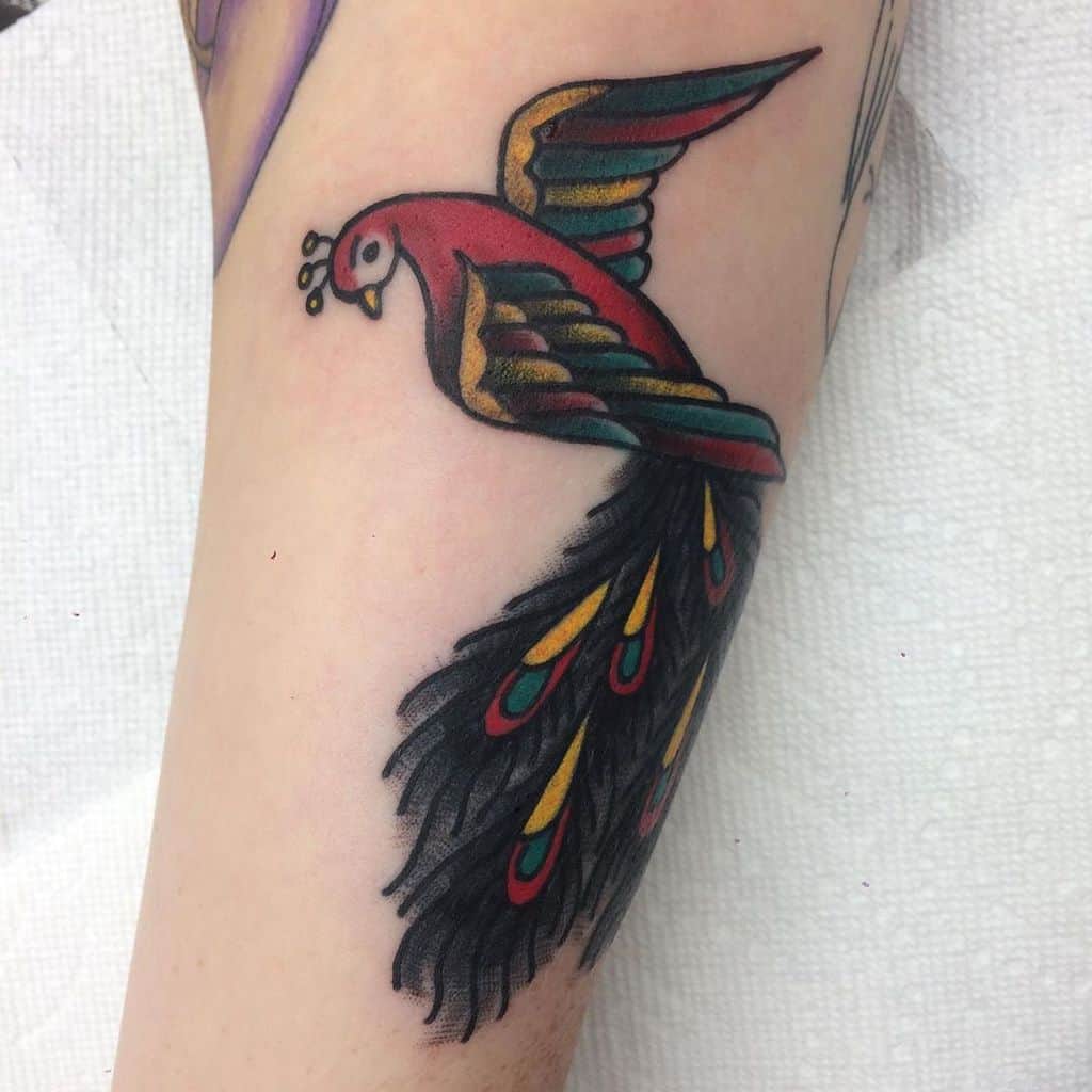 Traditional-style peacock tattoo on the forearm, featuring bold red, gold, and black tones with a flowing tail decorated with eye patterns.