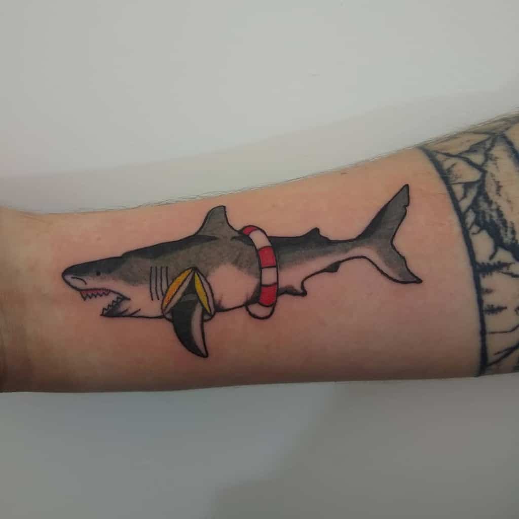A colorful tattoo of a shark wearing a red and white lifebuoy, blending humor with traditional tattoo styling on the forearm.