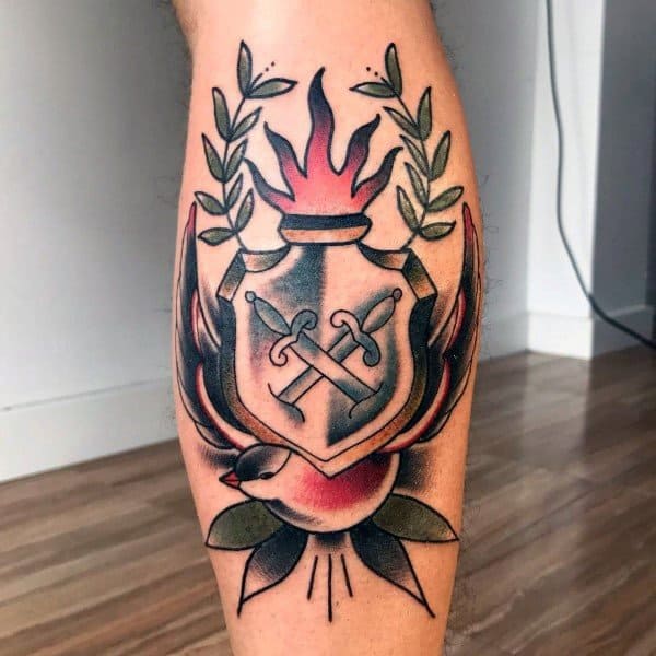 Colorful leg tattoo of a traditional-style shield with crossed swords, surrounded by laurel branches, flames, and a bird holding a leaf at the bottom.