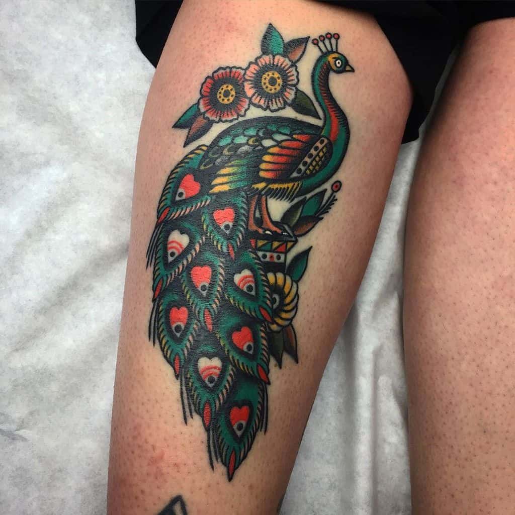 Vibrant peacock tattoo with colorful feathers in shades of green, blue, and red, accented with heart shapes and floral details on the thigh.