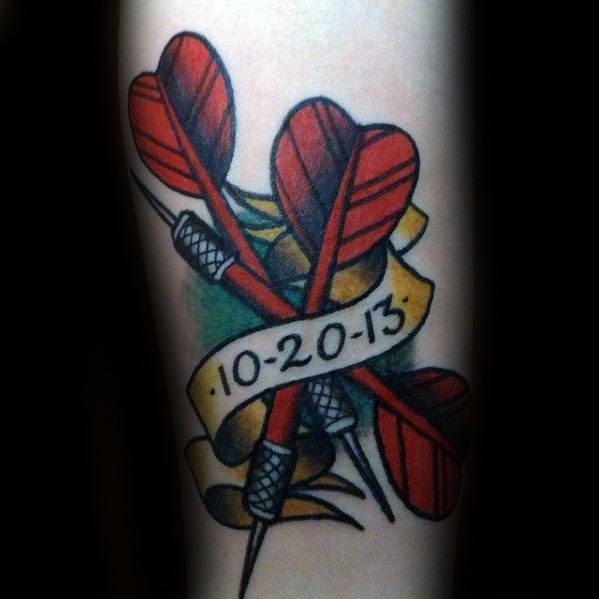 Colorful tattoo of three red-feathered darts crossed behind a banner with the date 10-20-13, symbolizing a meaningful and personal milestone.