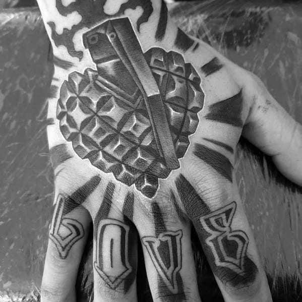 Black and grey 3D tattoo of a grenade-shaped heart on the hand, symbolizing explosive emotions, with "LOVE" inked across the fingers for contrast.