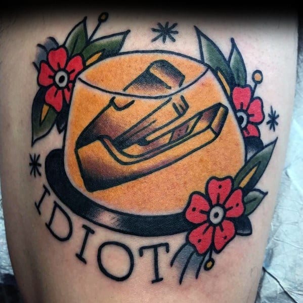 Color tattoo of a stapler encased in Jell-O, surrounded by flowers, with the word "IDIOT" below, referencing Jim’s classic prank on Dwight from *The Office*.