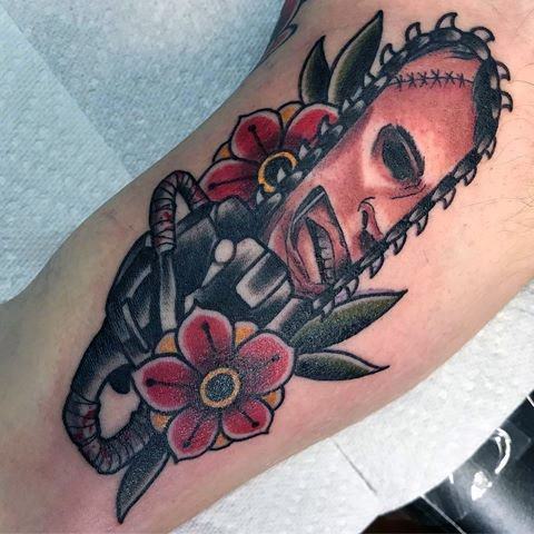 Neo-traditional tattoo of Leatherface’s stitched mask on a chainsaw, accented with bold flowers and vibrant colors for a unique horror-inspired design.