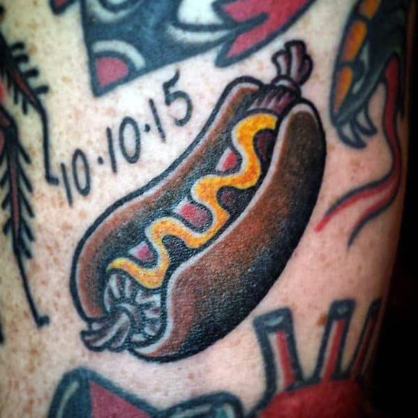 Tattoo of a hot dog with mustard, along with the date "10/10/15" above it, on the upper arm.