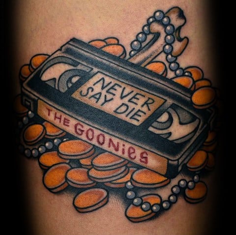Colorful tattoo of a VHS tape labeled "The Goonies" and "Never Say Die," surrounded by gold coins and pearls, inspired by the movie’s treasure theme.