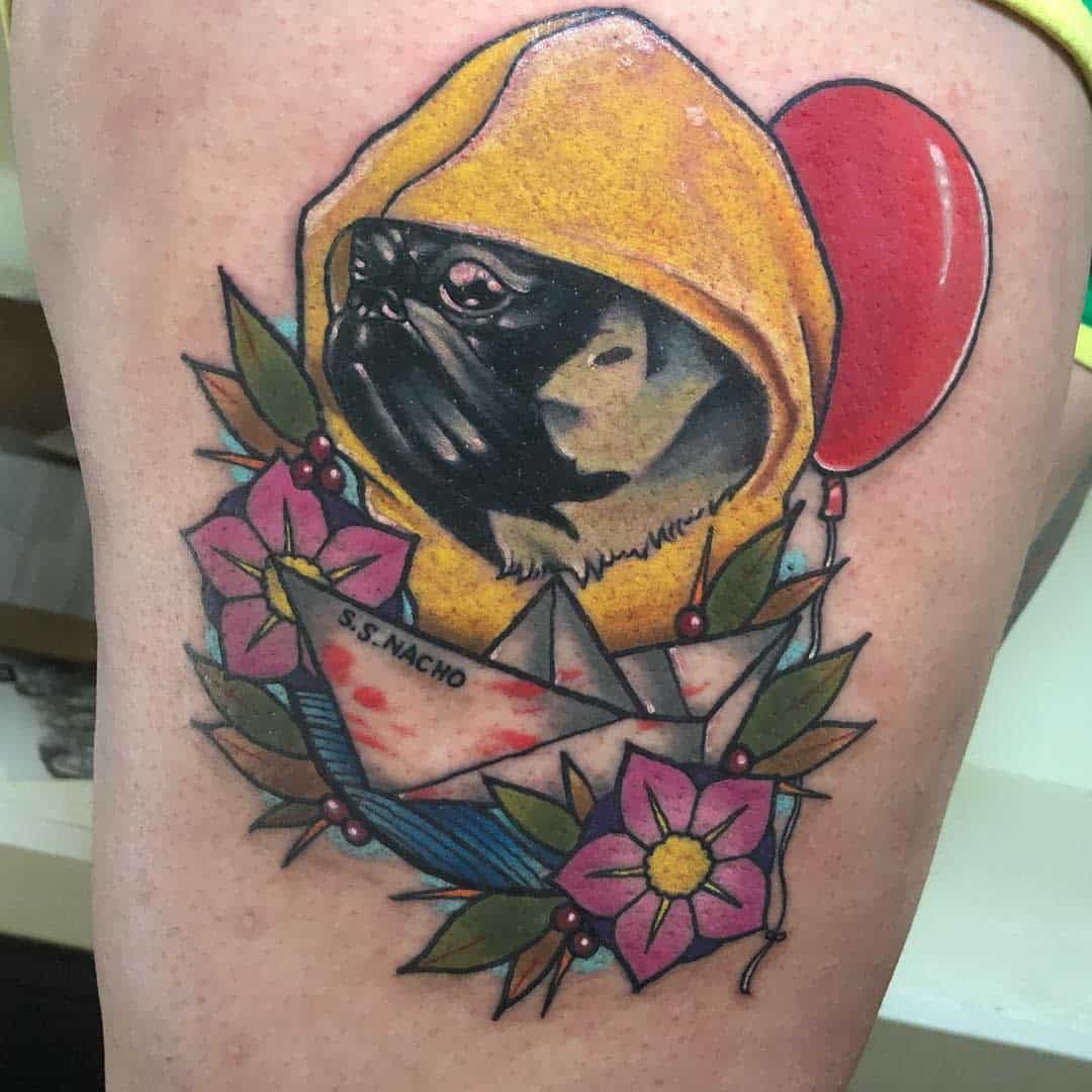 Tattoo of a pug in a yellow hood, red balloon, and paper boat with "S.S. NACHO," surrounded by flowers, berries and leaves