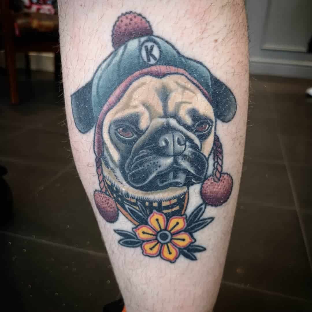 Tattoo of a pug wearing a winter hat with a pom-pom with letter 'K', a yellow checkered scarf, and a yellow flower with leaves below the chin