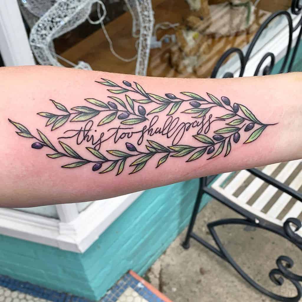 Tattoo with "this too shall pass" in cursive, surrounded by an olive branch design on the forearm.