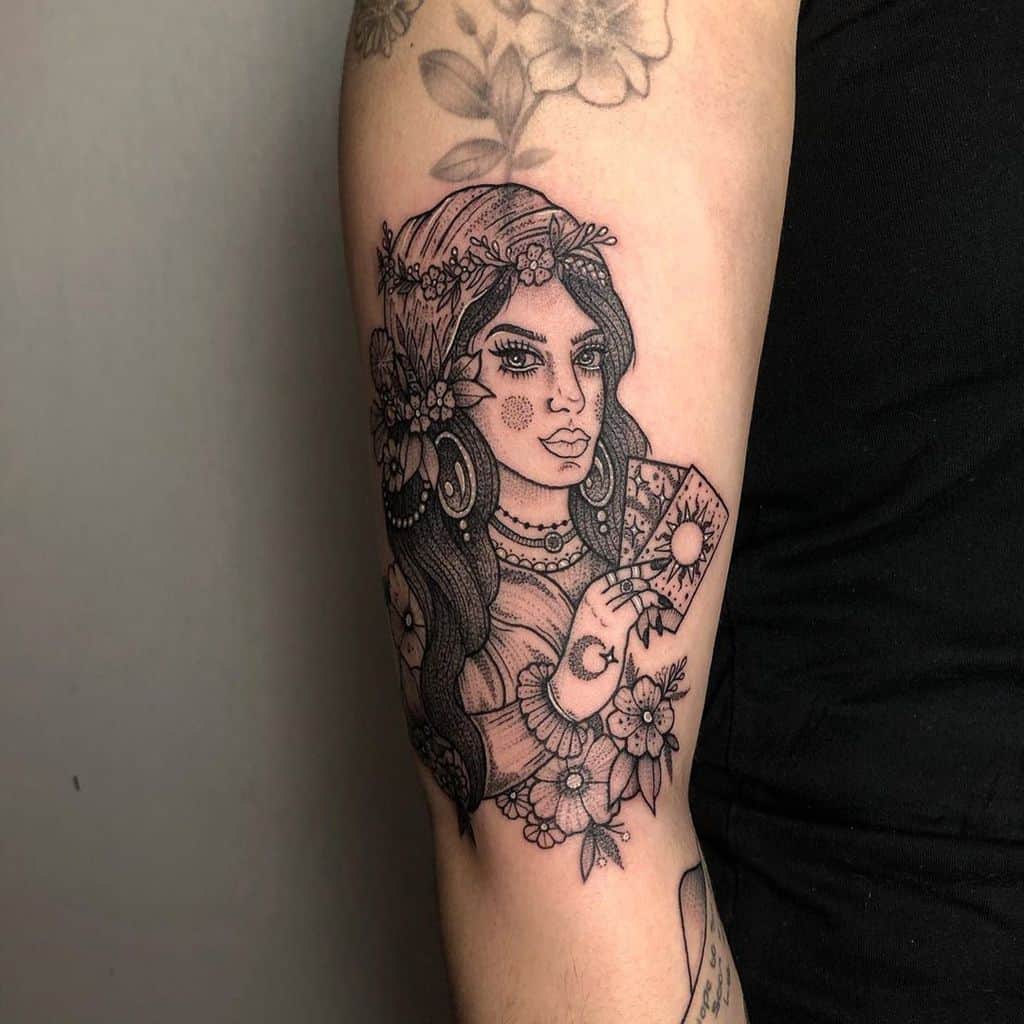 Black and grey tattoo of a mystical gypsy woman holding tarot cards, adorned with floral details, jewelry, and fine line shading on the upper arm.