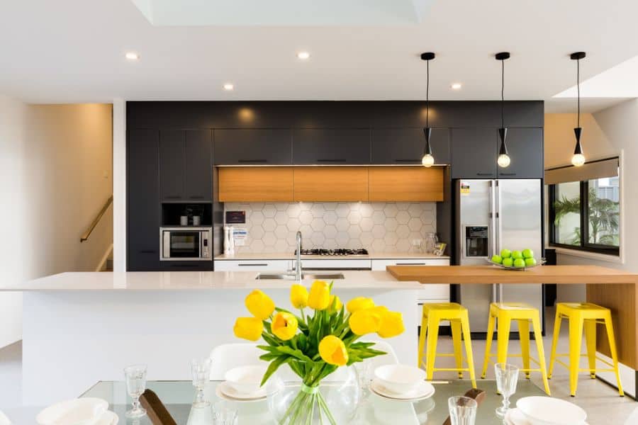 Modern kitchen with black cabinets, wood accents, white countertops, yellow stools, and pendant lights.