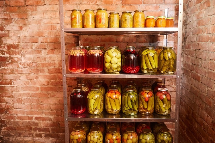 Metal shelf with jars of pickled vegetables and preserves against brick wall in basement.
