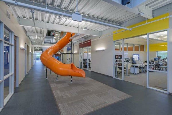 Office hallway with an orange slide and open-plan desks visible through sleek glass walls