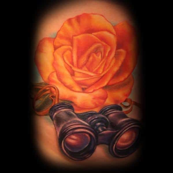 Realistic tattoo of black binoculars beside a vibrant orange rose, symbolizing observation, beauty, and adventure.