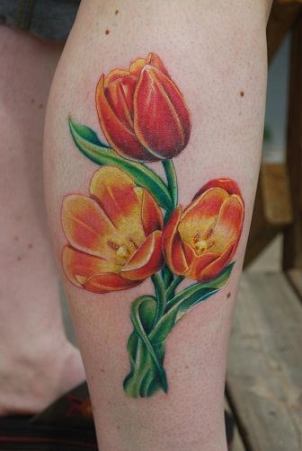 A vibrant tulip tattoo on the leg, featuring three orange and yellow flowers with rich green leaves, designed in a bold, realistic style.