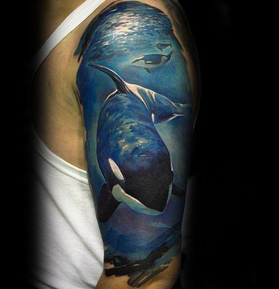 Realistic and colorful orca tattoo on the upper arm, showing whales swimming beneath water with detailed blue tones and shading.