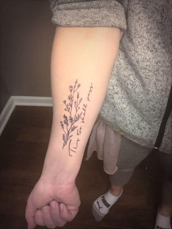 A graceful "This too shall pass" tattoo on the forearm, written in cursive alongside a delicate floral branch, symbolizing resilience and hope.