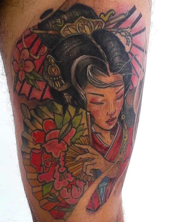 Vibrant Japanese-style tattoo of a Geisha holding a colorful fan, with intricate floral details, ornate hair accessories, and a rising sun backdrop.