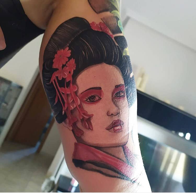 Vibrant tattoo of a Geisha with red floral hair ornaments, bold makeup, and a detailed kimono, blending realism with traditional Japanese art.