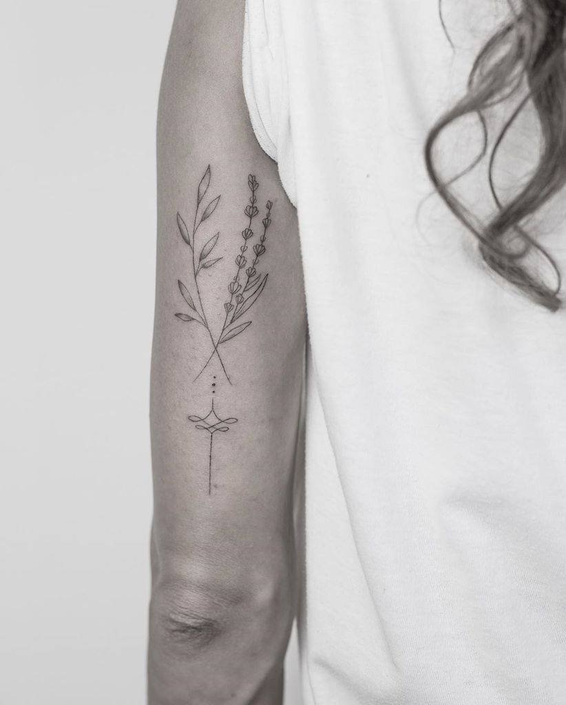 A minimalist tattoo of two vertical sprigs of lavender and leaves with delicate line details, accompanied by a central geometric design of dots and a cross beneath them, placed on the forearm.