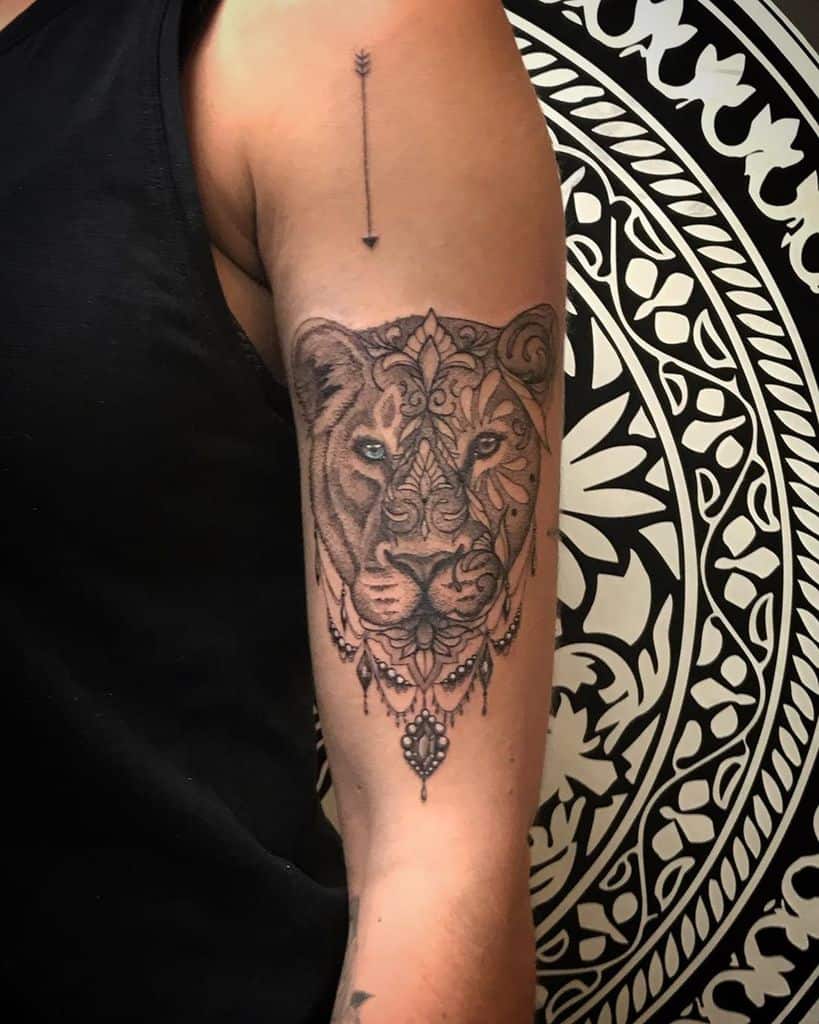 Intricate lioness tattoo on the upper arm, featuring mandala-inspired details, fine shading, and ornamental jewelry elements for a regal look.
