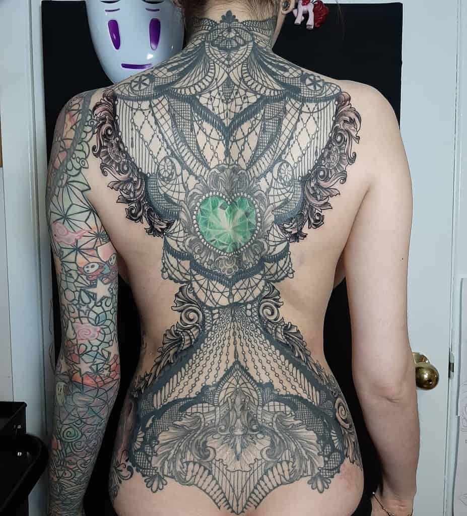 Full-back tattoo featuring an intricate lace corset design with ornate filigree, delicate beaded details, and a striking green gemstone centerpiece.