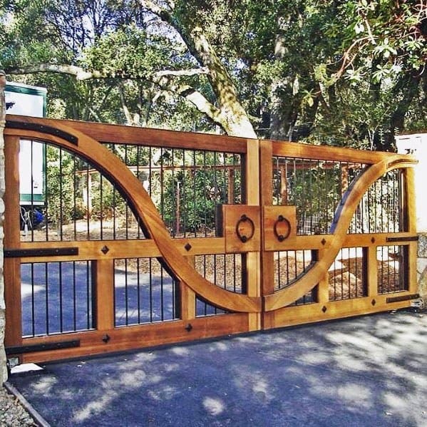 Ornate wooden and metal driveway gate with curved design, set against trees in a sunlit outdoor setting