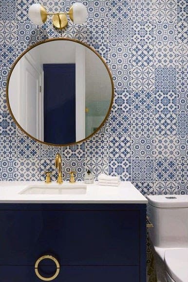 Bathroom with patterned blue and white tiles, navy blue vanity, gold faucet, and round mirror