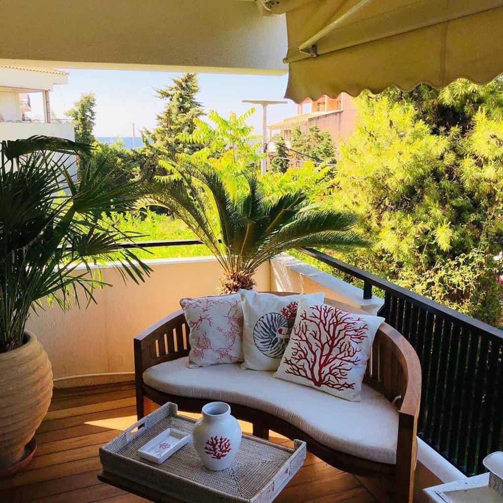 Cozy outdoor balcony with tropical plants, wooden seating, and coral-themed cushions.
