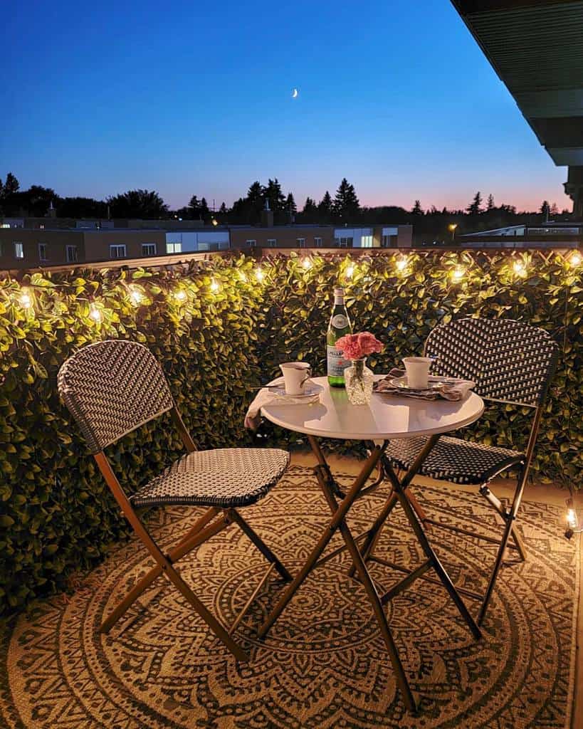 Intimate balcony at dusk with string lights, round table, chairs, and city view in the background.