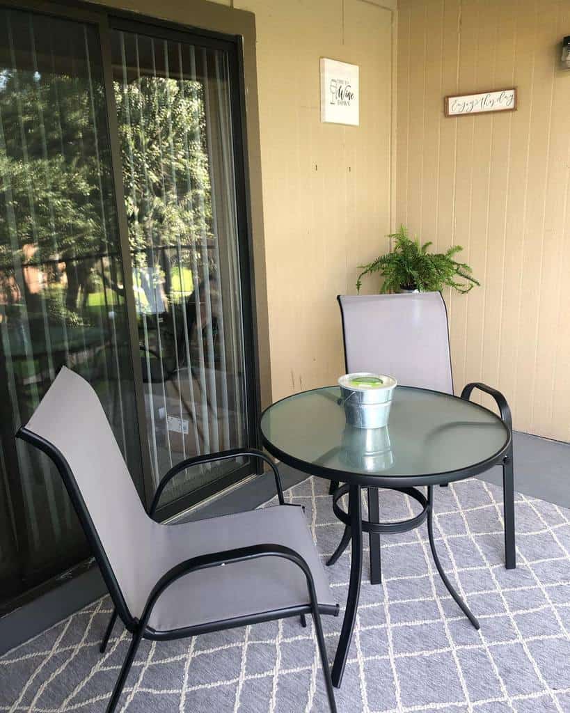 Simple patio with glass round table, two chairs, and a potted plant on a rug.