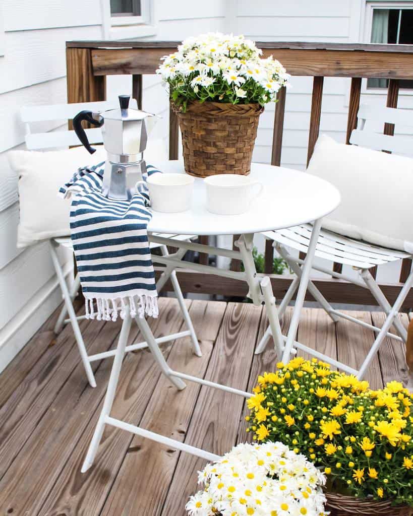Small patio with white folding furniture, daisy flowers, and a coffee setup on a wooden deck.
