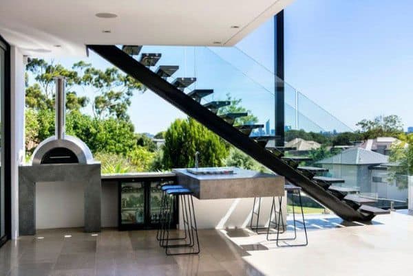 Sleek outdoor space featuring a modern pizza oven, bar seating, and a glass staircase balustrade.