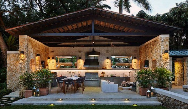 Expansive pavilion with a stone exterior, outdoor kitchen, dining area, and cozy lounge space surrounded by greenery.