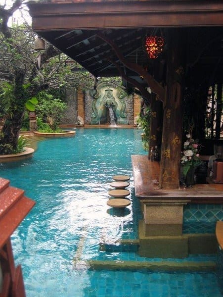 Poolside bar with round submerged stools, tropical decor, and ornate waterfall in the background.