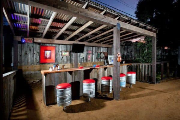 Rustic outdoor bar with keg stools, wooden countertops, and corrugated metal roof in an industrial style.