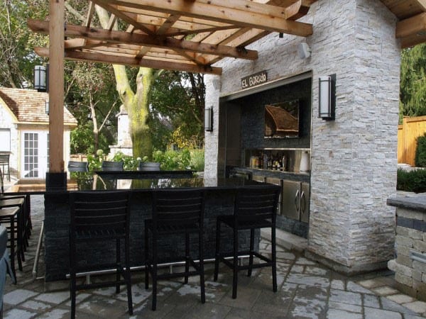 Modern backyard bar with stone walls, black countertops, and wooden pergola for shade.