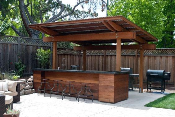 Wood pallet bar with metal stools and a corrugated roof in a shaded backyard setting.