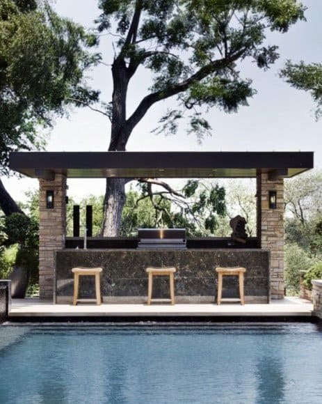 Contemporary poolside bar with stone columns, polished countertop, and wooden stools under a shelter.