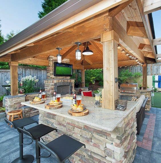 Rustic outdoor bar with stone countertops, wooden pavilion, and mounted TV, surrounded by stools.