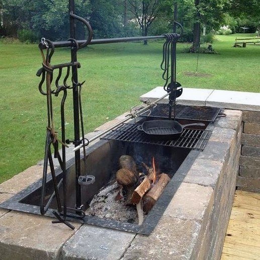 Outdoor stone fireplace with metal grill setup, featuring a skillet and burning logs in a lush, grassy yard