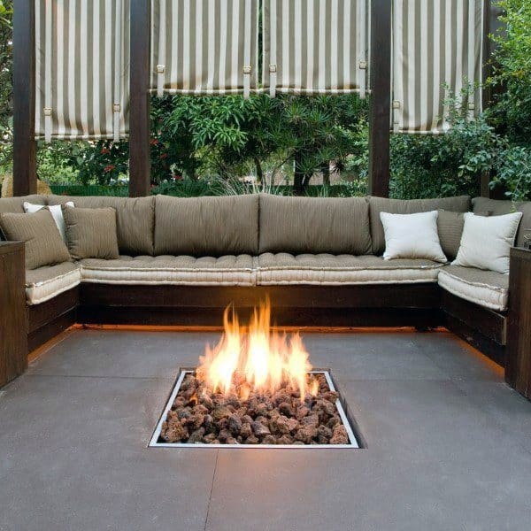 Cozy outdoor seating features a beige sectional, striped curtains, and a fire pit glowing warmly in the center