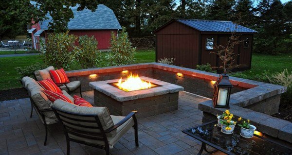 Cozy patio with a central fire pit, surrounded by chairs, next to a dark wooden shed and trees